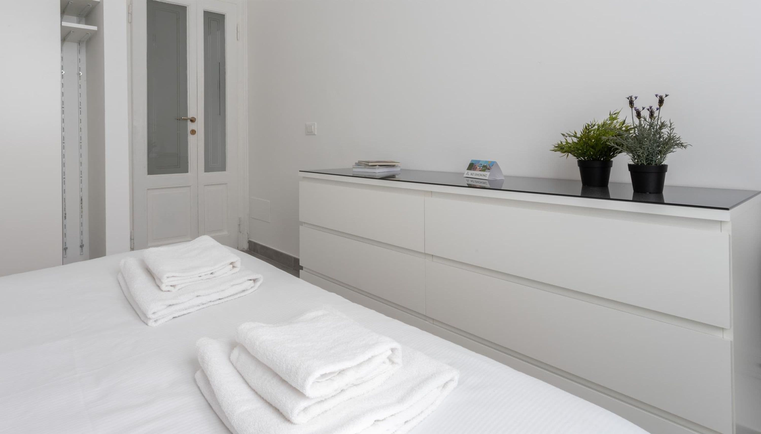 A modern hotel room with a bed, a white wardrobe, and a small shelf with plants and a no-smoking sign.