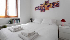 A neatly made bed with white linens and towels, a bedside table with a lamp, a bookshelf with books, and a colorful abstract painting on the wall. - Italianway Comfort - Villoresi 25 (Milan)