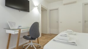 Une chambre d’hôtel équipée d’un téléviseur à écran plat fixé au mur, d’un bureau avec un ordinateur portable, d’un fauteuil de bureau gris, d’un lit avec du linge de lit blanc et d’une armoire blanche.