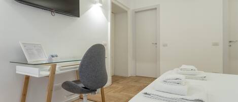 Hotellihuone, jossa on seinälle asennettu taulutelevisio, työpöytä kannettavalla tietokoneella, harmaa toimistotuoli, valkoisilla lakanoiden peitetty sänky ja valkoinen vaatekaappi.