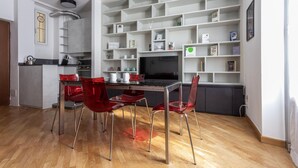 A modern kitchen with a dining area, featuring a black table, red chairs, and a white shelving unit with various items. - Italianway Easy - Abruzzi 19 (Milan)