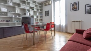 A modern living room with a dining area, featuring a glass table, red chairs, a red sofa, and a white shelving unit with various items. - Italianway Easy - Abruzzi 19 (Milan)