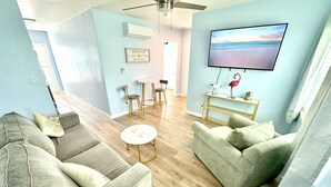 Een woonkamer met een plafondventilator, een aan de muur bevestigde televisie, een eethoek met een tafel en stoelen, en een lichtblauwe muur.