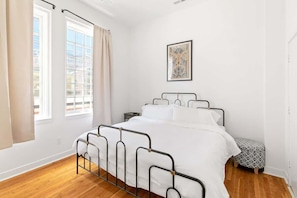 A bedroom with a metal bed frame, white bedding, a small bedside table, a patterned ottoman, and a window with curtains.