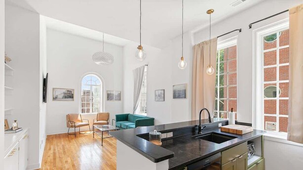 Et moderne køkken med en central ø, et spiseområde med en grøn sofa, og udsigt til en murstensbygning gennem store vinduer.