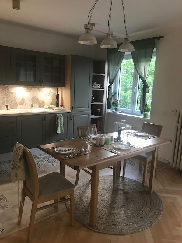 Comedor con mesa de madera dispuesta para cuatro, cocina al fondo y ventana con cortinas.