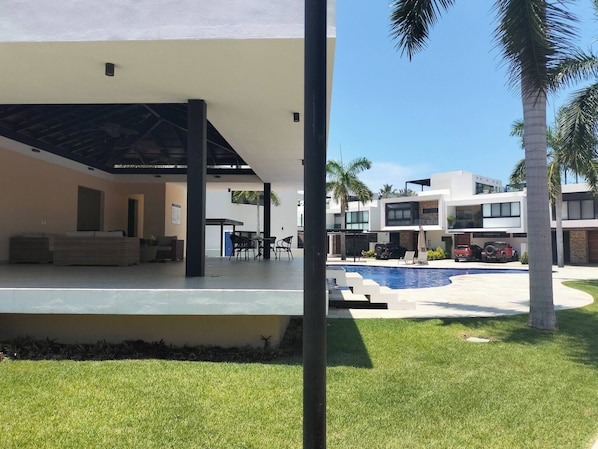 A modern building with a covered outdoor seating area, a swimming pool, and palm trees.