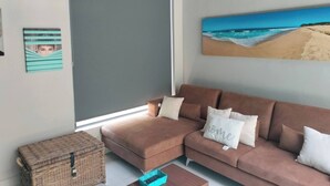 Een woonkamer met een bruine bank, een rieten kist en een schilderij van een strand aan de muur.