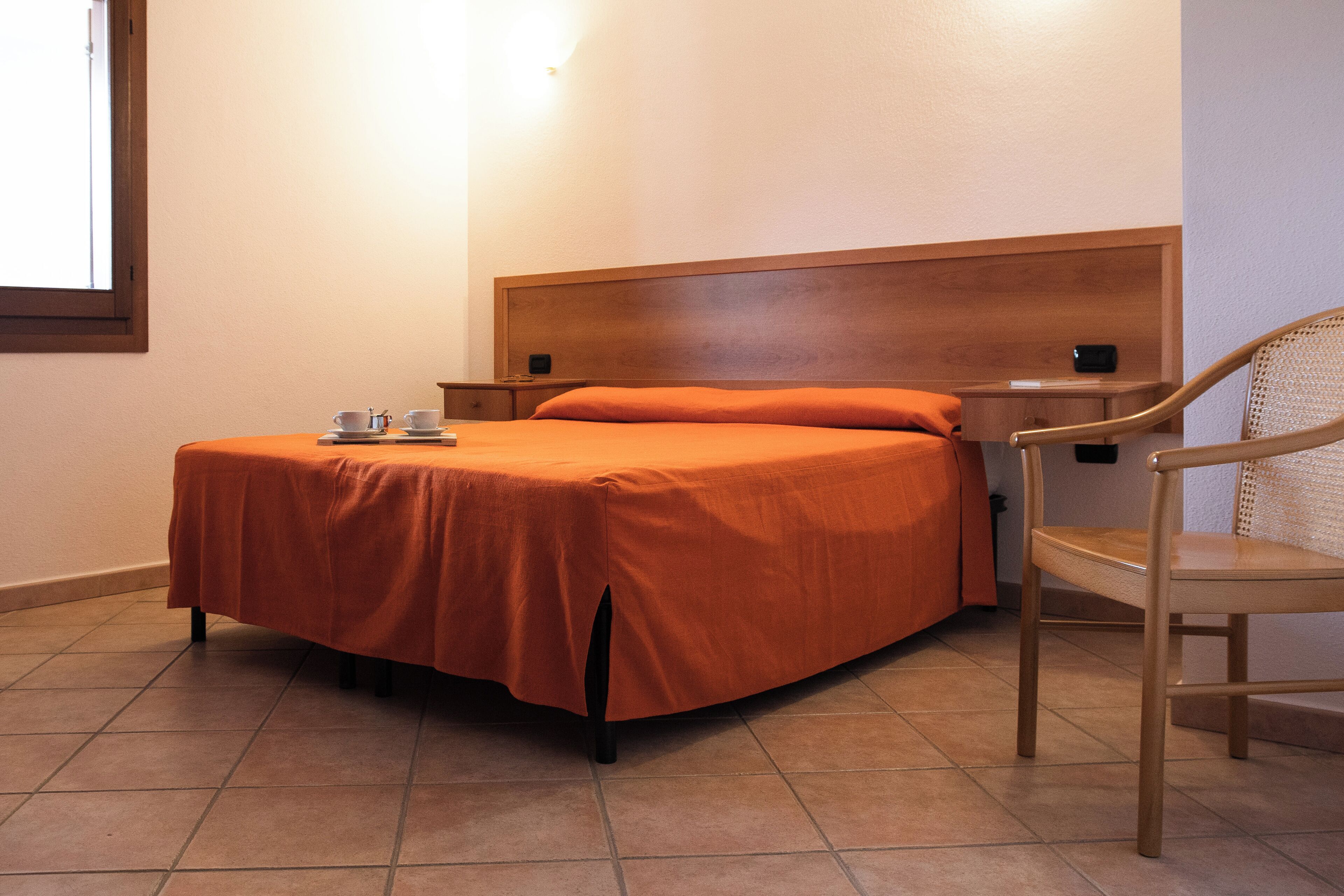 A single bed with an orange bedspread, a wooden headboard, a nightstand with a phone, and a chair.