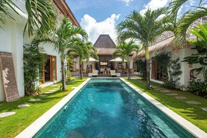 A swimming pool with a thatched-roof building in the background, surrounded by palm trees and a well-maintained lawn. - Villa Parasol by Alfred in Bali (Seminyak)