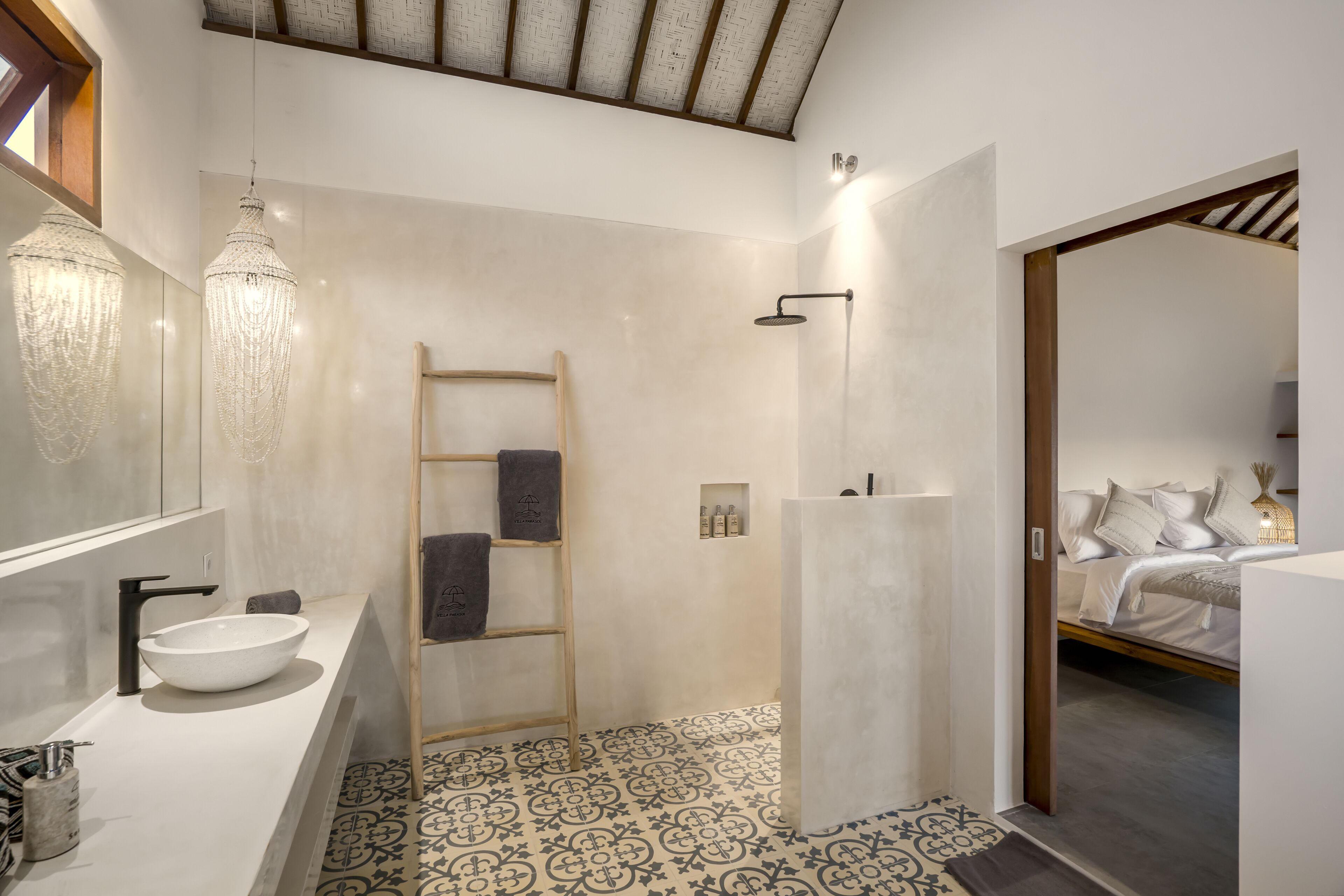 A modern bathroom with a white sink, a wooden ladder, a shower area, and a bed visible through an open door.