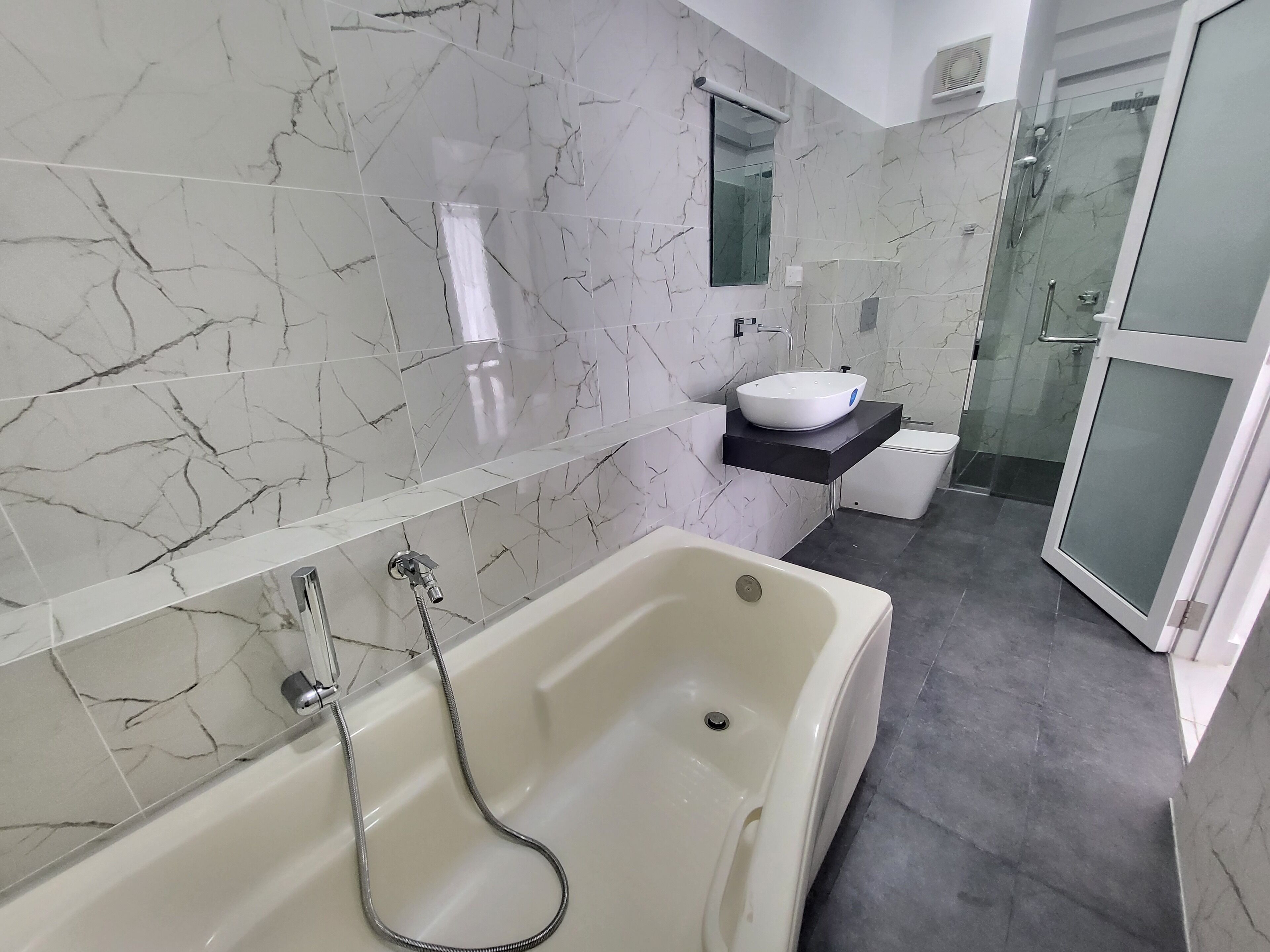Un baño moderno con pared revestida de mármol, una bañera independiente, ducha con cerramiento de vidrio y un lavamanos pequeño con encimera negra.