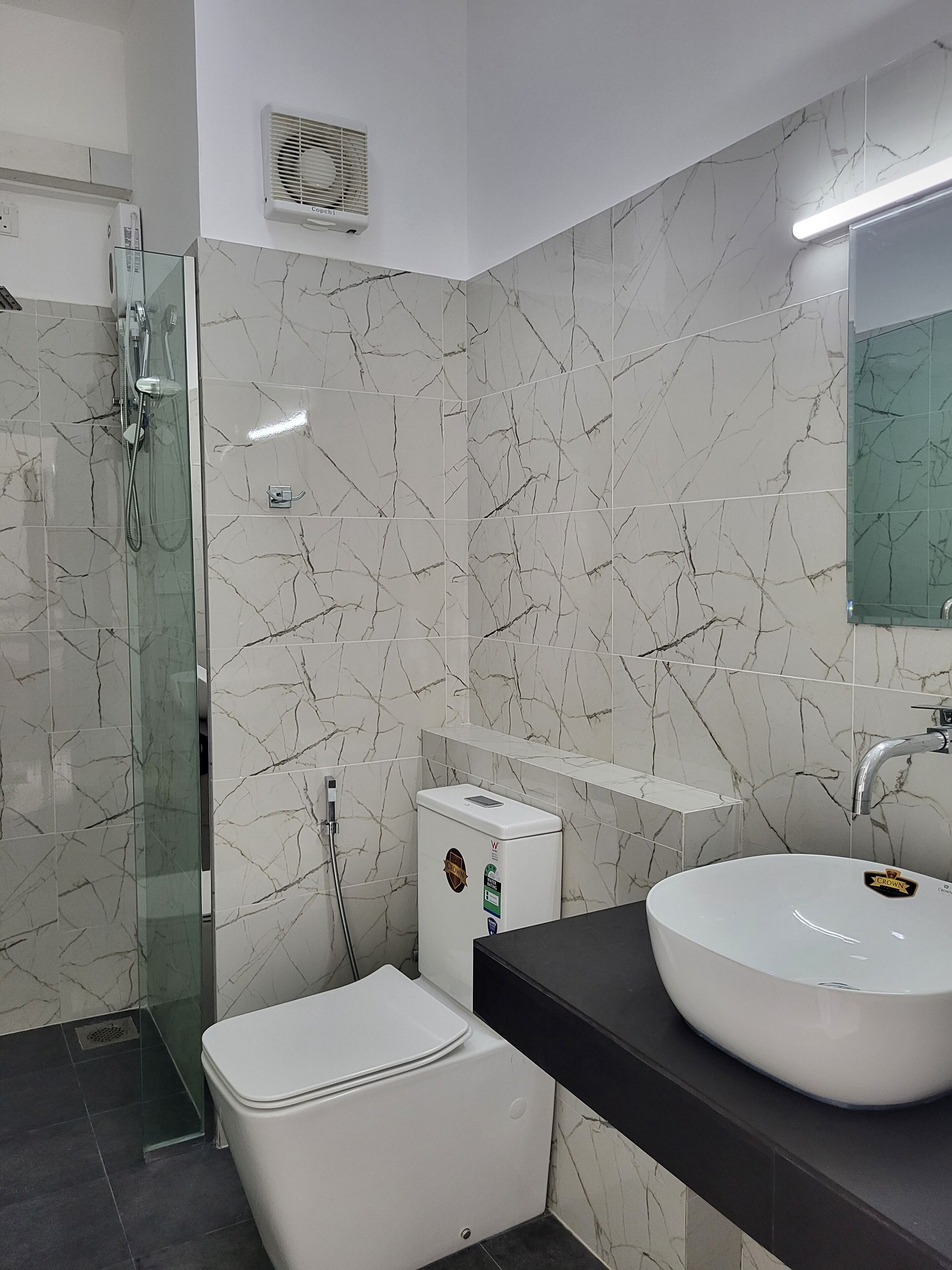 A modern bathroom with a glass-enclosed shower, a white toilet, a white sink with a mirror above it, and a marble-patterned wall.