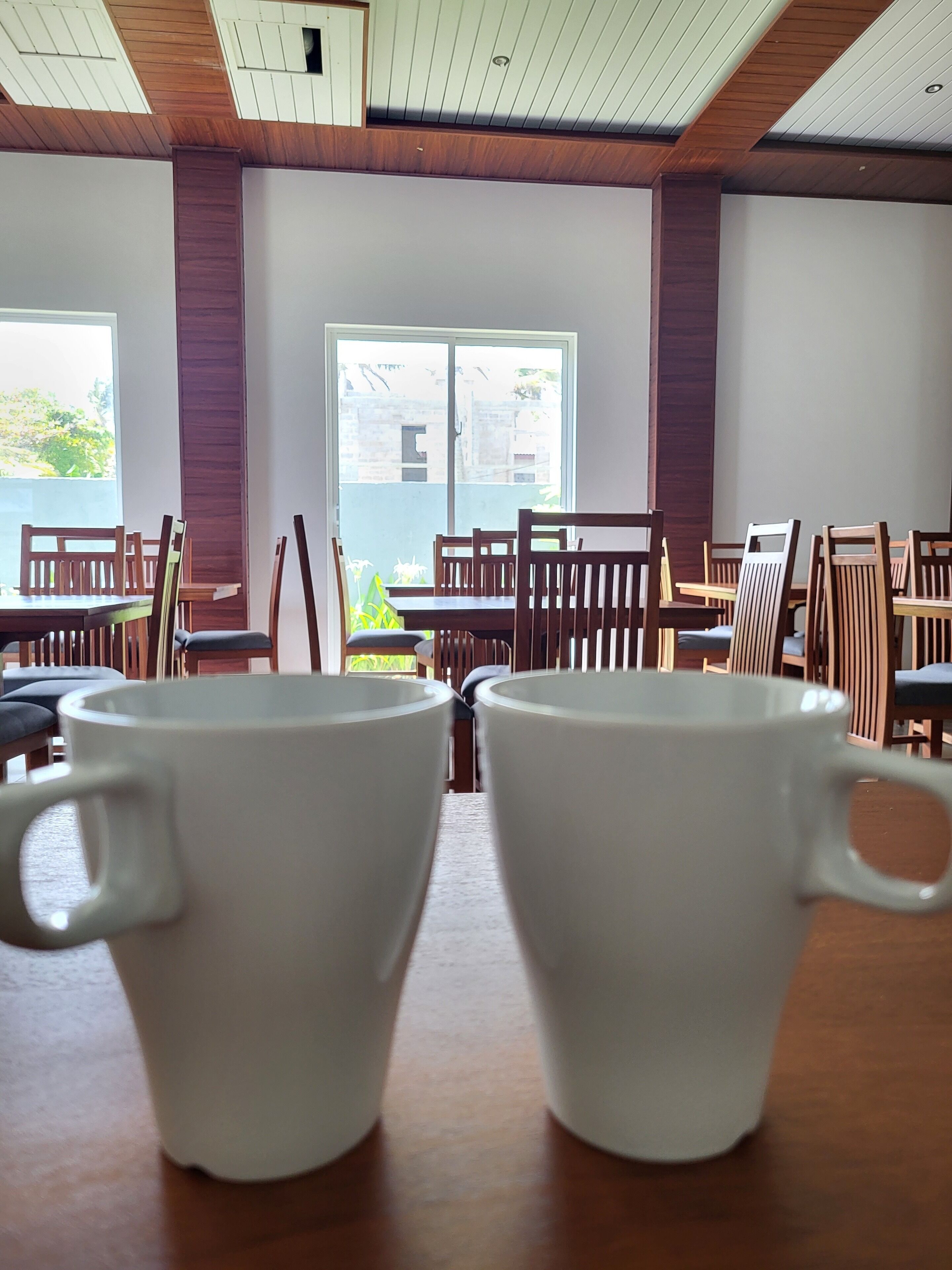 Un comedor con mesas y sillas de madera, dos tazas blancas sobre una mesa y un ventanal con vistas a la vegetación.