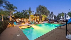 A swimming pool with lounge chairs and a slide, surrounded by trees and a building with outdoor lighting. - Resort-Style 5 BR Retreat in Lake Hodges W Pool, Jacuzzi, Grill, Volleyball Court (Escondido)