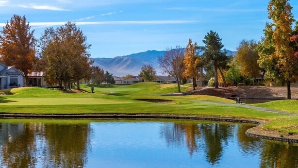 Campo de golf con un estanque, casas y montañas al fondo.