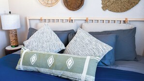 Una cama con colcha azul, dos almohadas y un cojín decorativo con diseño tejido. Hay cestas tejidas en la pared detrás de la cama.