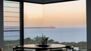 A corner room with a large window overlooking a body of water and a distant landmass, a round table with chairs, and a small plant on the table. - 'Cloud Nine' - Spa, Sauna, Iconic Jervis Bay Views (Vincentia)