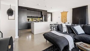 A modern living room with a dark sofa, a coffee table, and a kitchen area with a central island. - 'Cloud Nine' - Spa, Sauna, Iconic Jervis Bay Views (Vincentia)