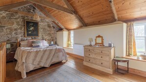 A cozy bedroom with a stone wall, wooden ceiling beams, a bed with a floral bedspread, a dresser, and a window with curtains.