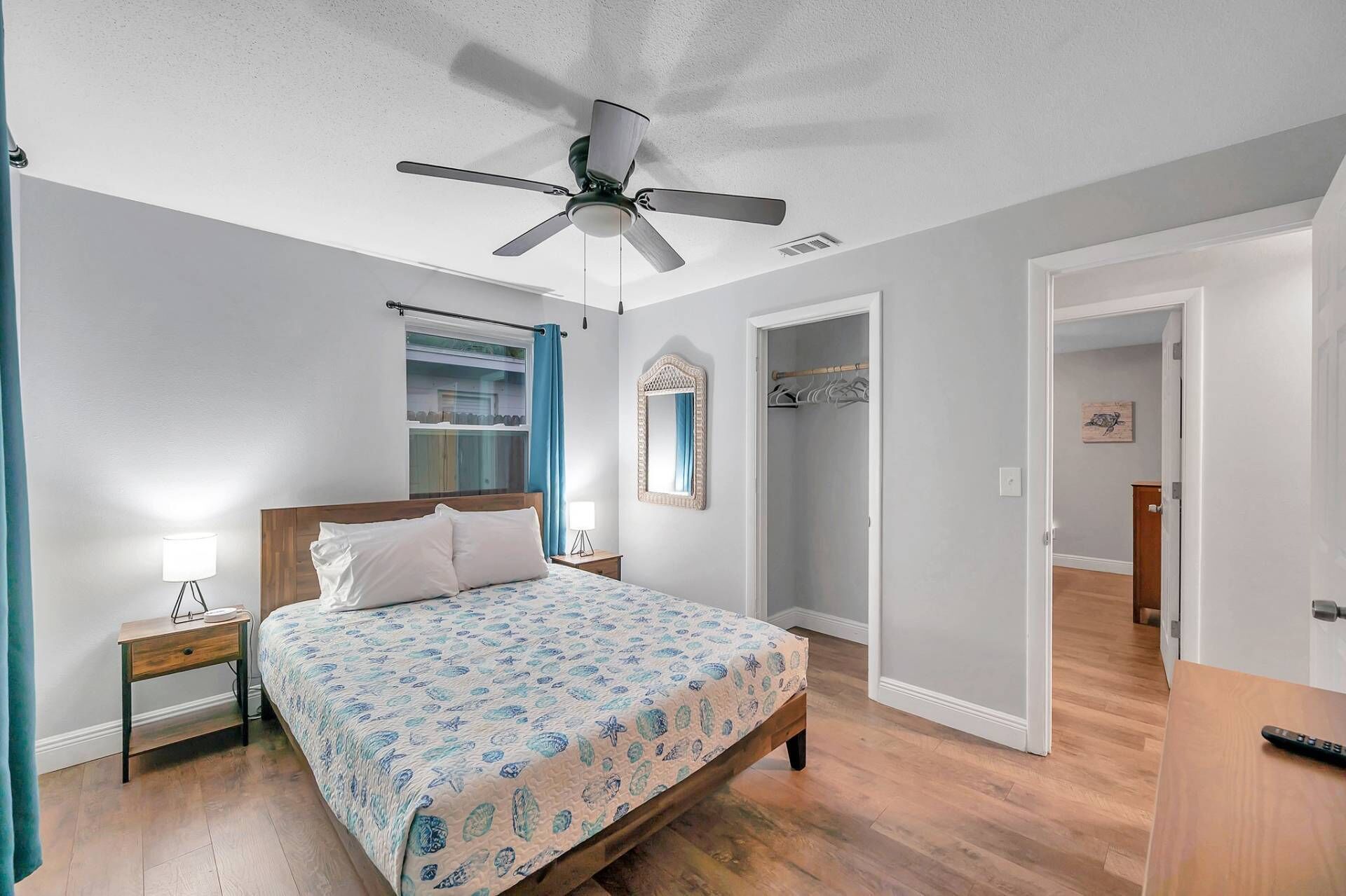 Un dormitorio con cama, mesita de noche, ventilador de techo, ventana con cortinas, espejo y un pasillo que lleva a otra habitación.