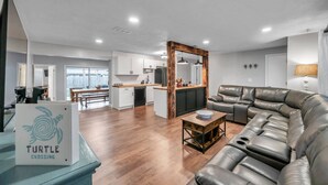 A modern living room with a leather sofa, a wooden coffee table, and a kitchen area with white cabinets and a wooden accent wall. - Quiet Neighborhood! Close to Beach. Private Backyard w/ Gas Grill ~ Turtle Crossing (Largo)