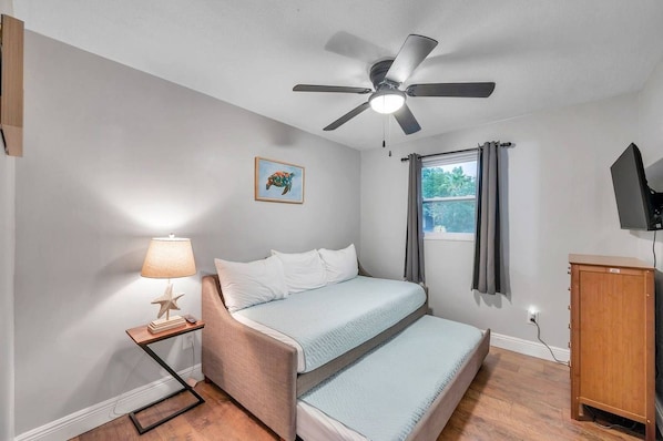 A bedroom with a bed, a ceiling fan, a window with curtains, a bedside lamp, and a small wooden cabinet.