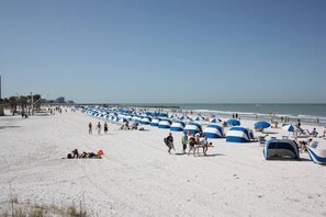 Une plage parsemée de nombreux parasols et tentes bleus et blancs, un rivage sablonneux et un ciel dégagé.