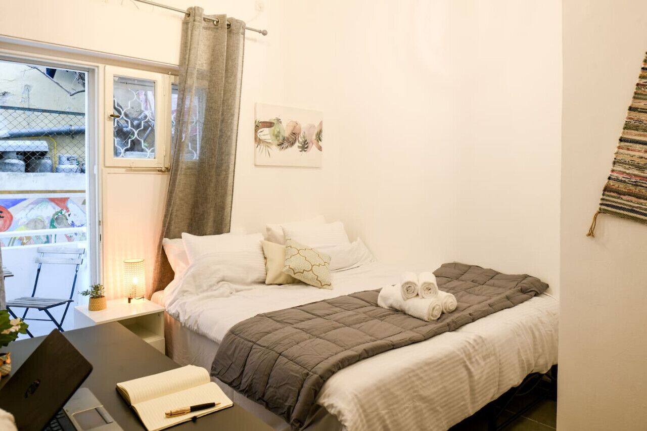 A bedroom with a bed, a desk with a laptop and a notebook, a window with a curtain, and a wall-mounted artwork.