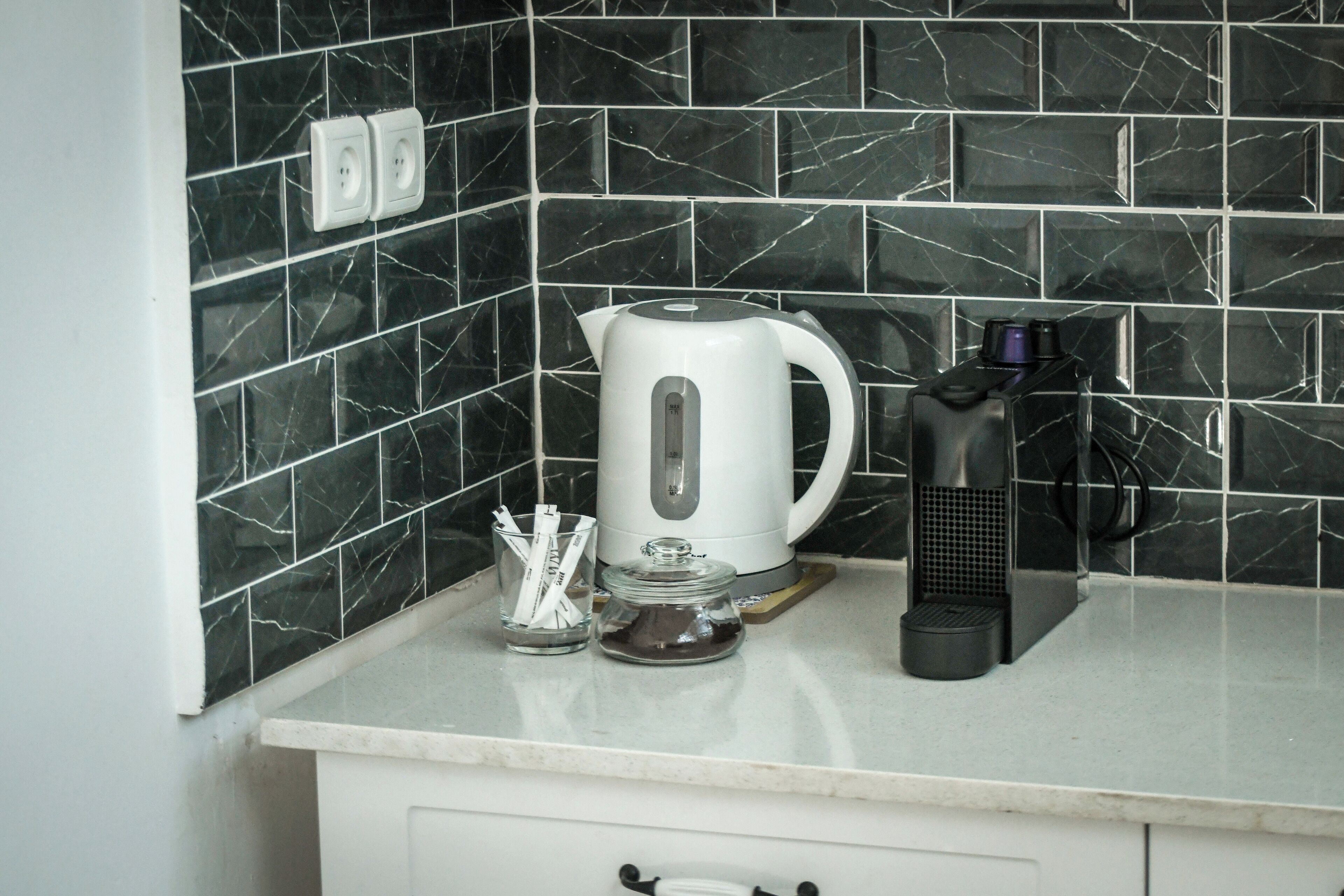 Una cocina con salpicadero de mármol, un hervidor blanco, una cafetera y un recipiente de vidrio con utensilios.