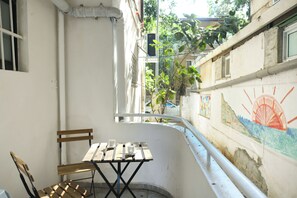 A narrow balcony with a wooden table and chairs, a white wall with a mural, and a view of the street and greenery. - Charming 2BR in Haifa by Sea N' Rent (Haifa)