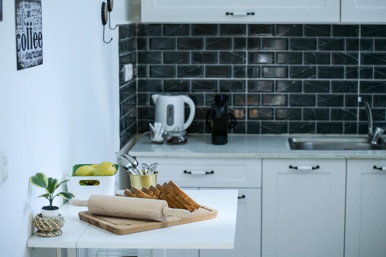 Eine moderne Küche mit weißen Schränken, einer schwarzen Fliesenrückwand, einem Wasserkocher und einem Schneidebrett mit Brot.