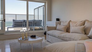 Un soggiorno moderno con una grande finestra vista mare, un divano beige, un tavolino rotondo con candele e un balcone con posti a sedere all'aperto.