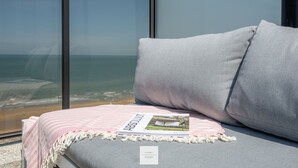 Un divano grigio con una rivista sopra, una coperta a righe rosa e bianca, e vista sulla spiaggia attraverso ampie finestre.