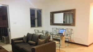 Ein Wohnzimmer mit einer Couch, einem Couchtisch und einem auf einem Ständer stehenden Laptop. Ein Fenster mit Metallrahmen und ein Spiegel sind an der Wand zu sehen.