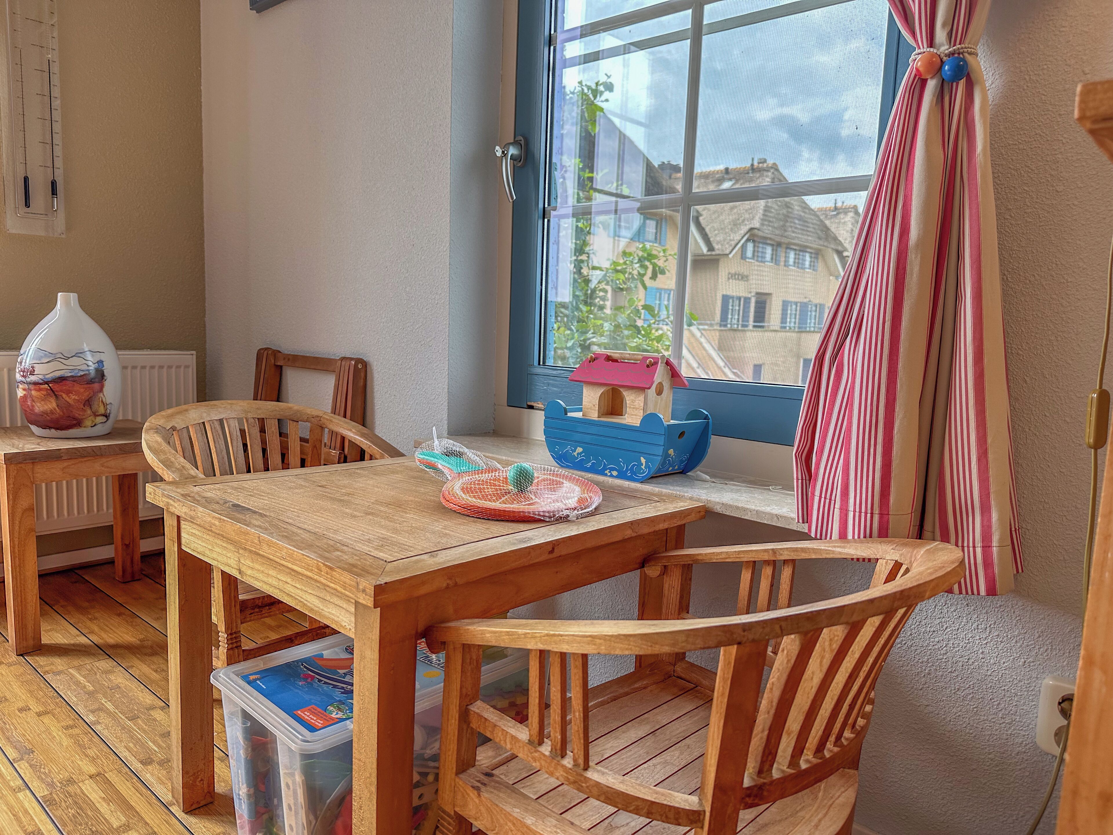 A cozy room with a wooden table and chairs, a window with a view of buildings, a small wooden boat on the windowsill, and a vase on a side table.