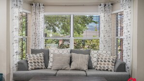 Un canapé gris agrémenté de coussins à motifs, une table basse en bois et une fenêtre avec des stores blancs et des rideaux à fleurs.