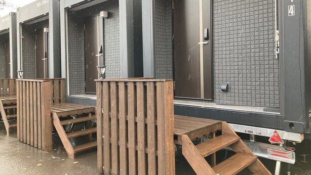Una fila de baños portátiles con estructuras metálicas y paredes cubiertas de azulejos negros. Cada cabina tiene un banco de madera en el exterior.