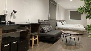 Habitación de hotel moderna con sofá, escritorio pequeño con lámpara, cama y planta en maceta.