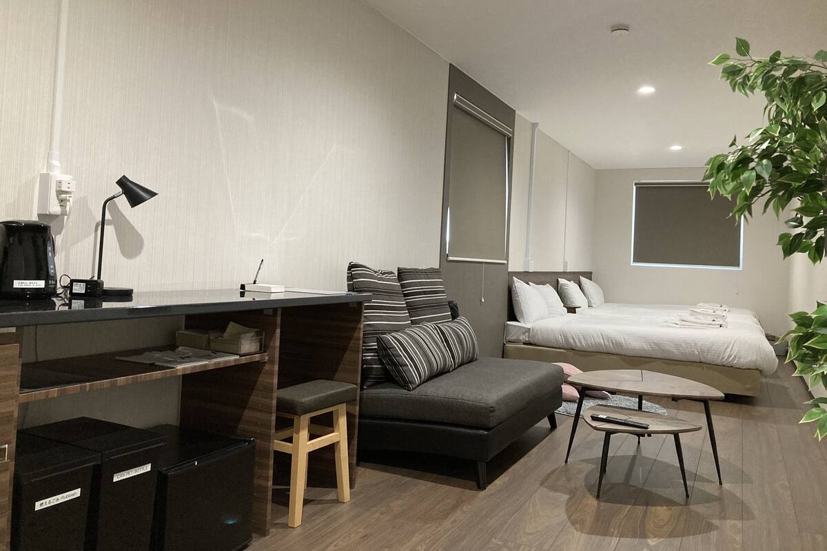 Habitación de hotel moderna con sofá, escritorio pequeño con lámpara, cama y planta en maceta.