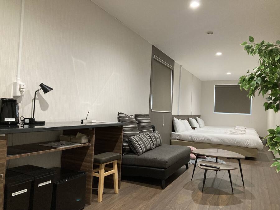 A modern hotel room with a sofa, a small desk with a lamp, a bed, and a potted plant.