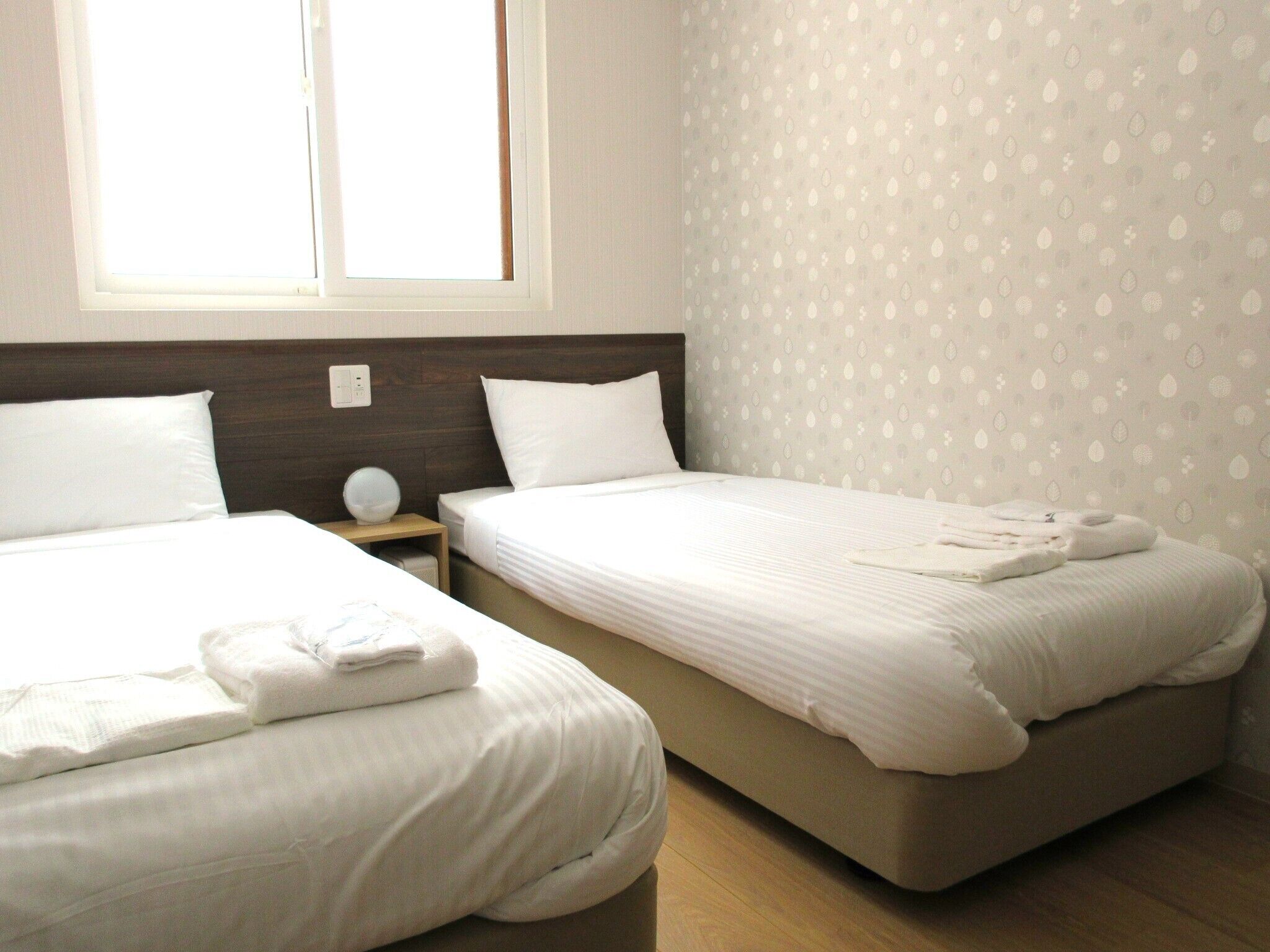 A hotel room with two beds, a wooden headboard, a nightstand with a lamp, and a window with blinds.