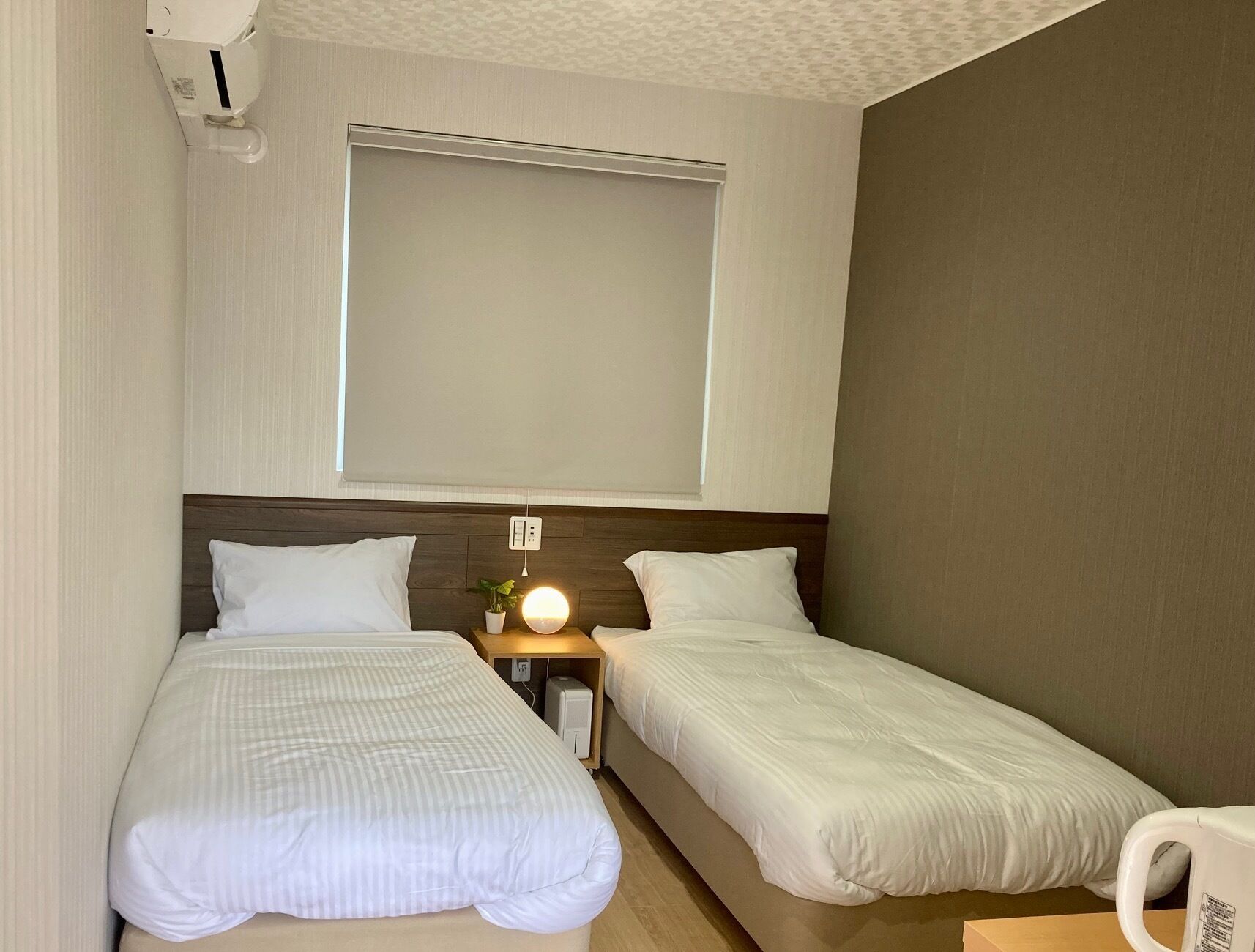 A hotel room with two single beds, a bedside table with a lamp, a power outlet, and a window with blinds.