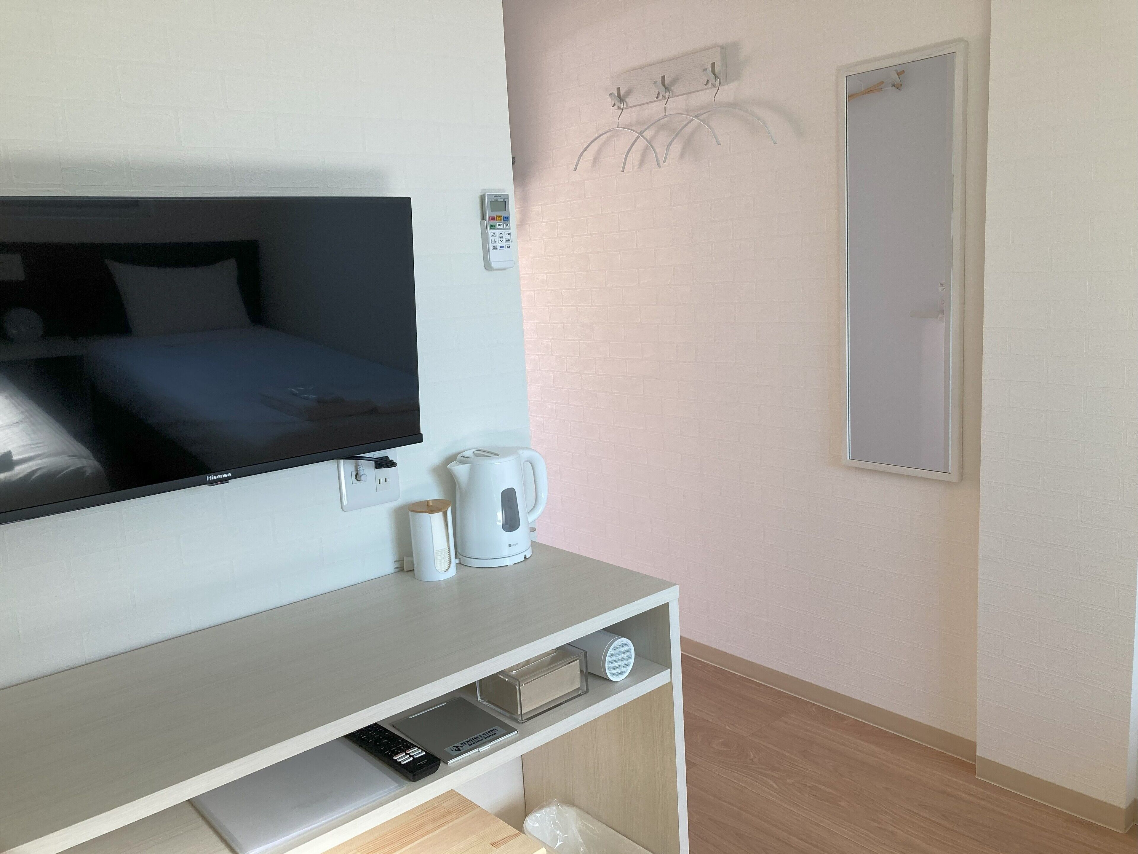 A hotel room with a bed, a TV, a kettle, a coffee maker, a remote control, and a small shelf with a speaker and a book.