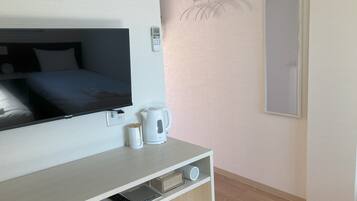 A hotel room with a bed, a TV, a kettle, a coffee maker, a remote control, and a small shelf with a speaker and a book.