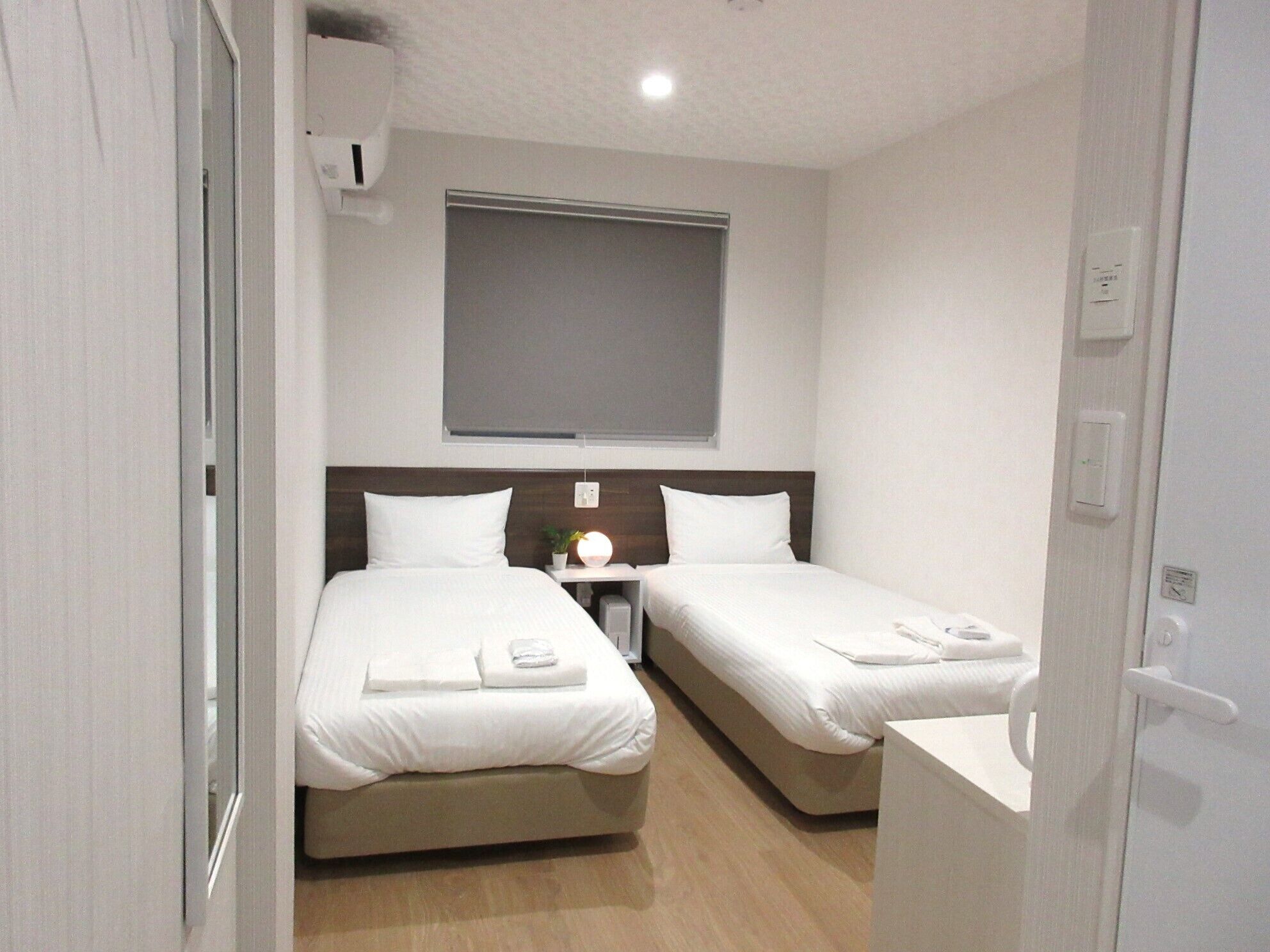 A hotel room with two single beds, a bedside table with a lamp, a window with a blind, and a wooden floor.