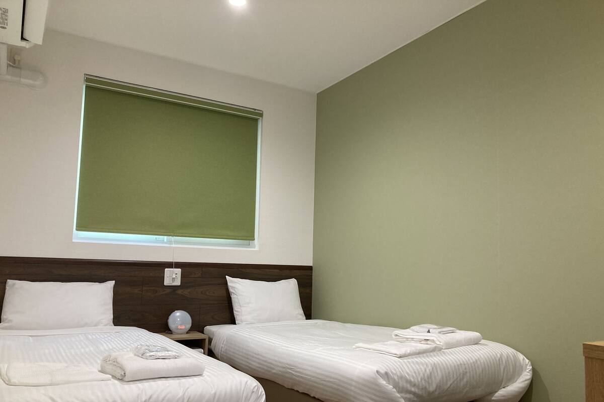 Una habitación de hotel con dos camas, una mesita de noche, un reloj en la pared y una ventana con persiana verde.