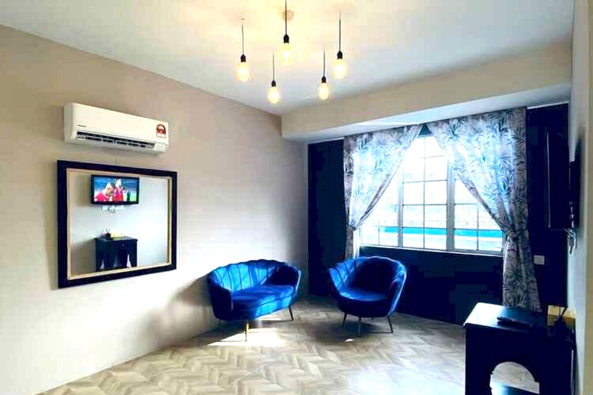 A spacious room with a herringbone floor pattern, two blue armchairs, a small table, and a TV mounted on the wall.