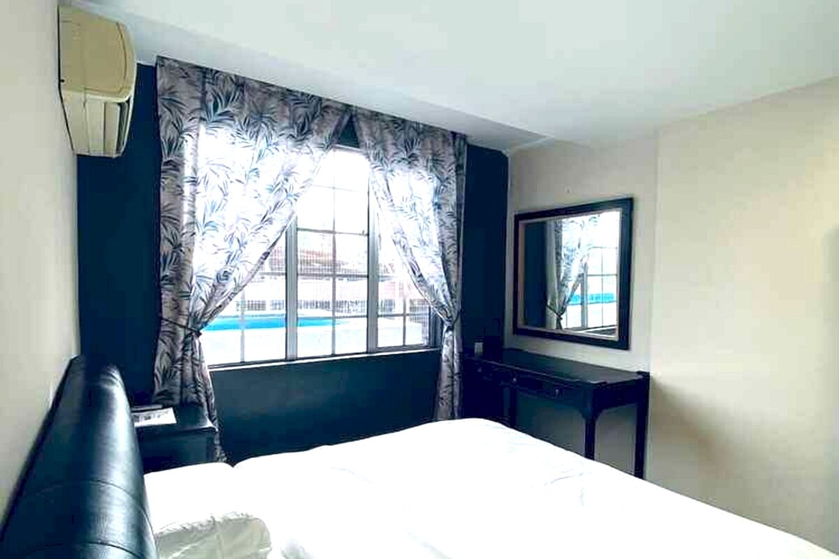 A hotel room with a large bed, a desk, a chair, a window with curtains, and a wall-mounted mirror.