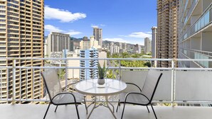 A balcony with a round table and two chairs, overlooking a cityscape with high-rise buildings. - 18th Fl Ilikai Hotel Condo | King Bed | Valet Parking | City View (Honolulu)